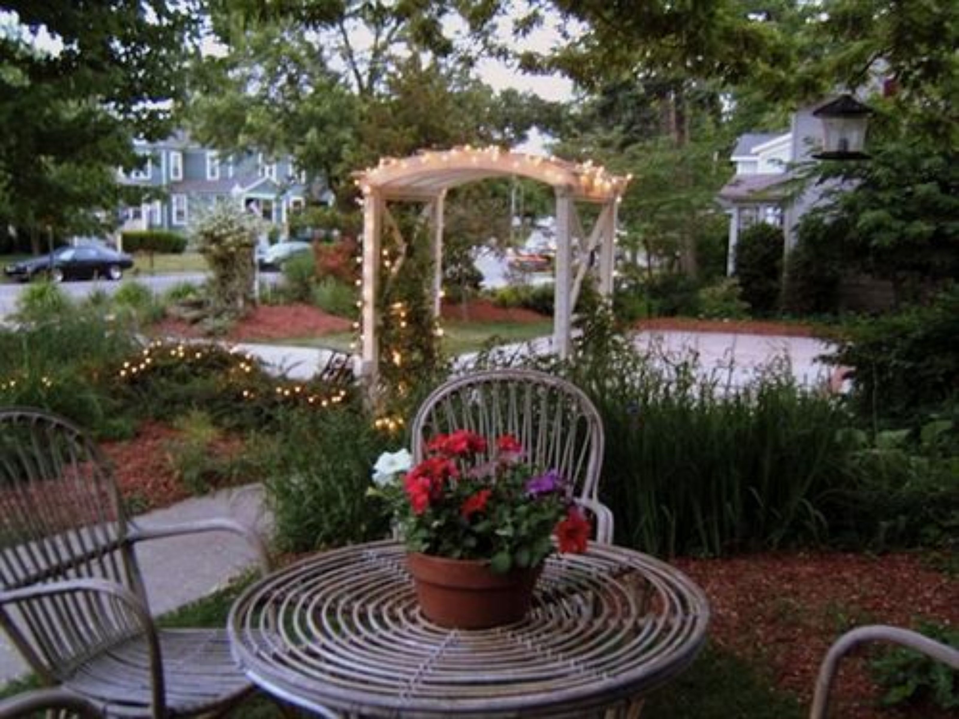Um jardim com mesa e cadeiras de metal, uma pérgula de madeira com luzes de cordão e um vaso com flores vermelhas.
