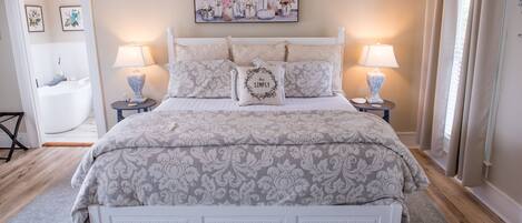 A neatly arranged bedroom with a bed, two bedside tables with lamps, a large mirror, and a painting on the wall.