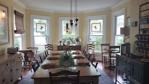 A dining area with a long table set for six, surrounded by chairs. There are windows with curtains, a chandelier, and kitchen appliances on the right. - Pecan Tree Inn (Beaufort)