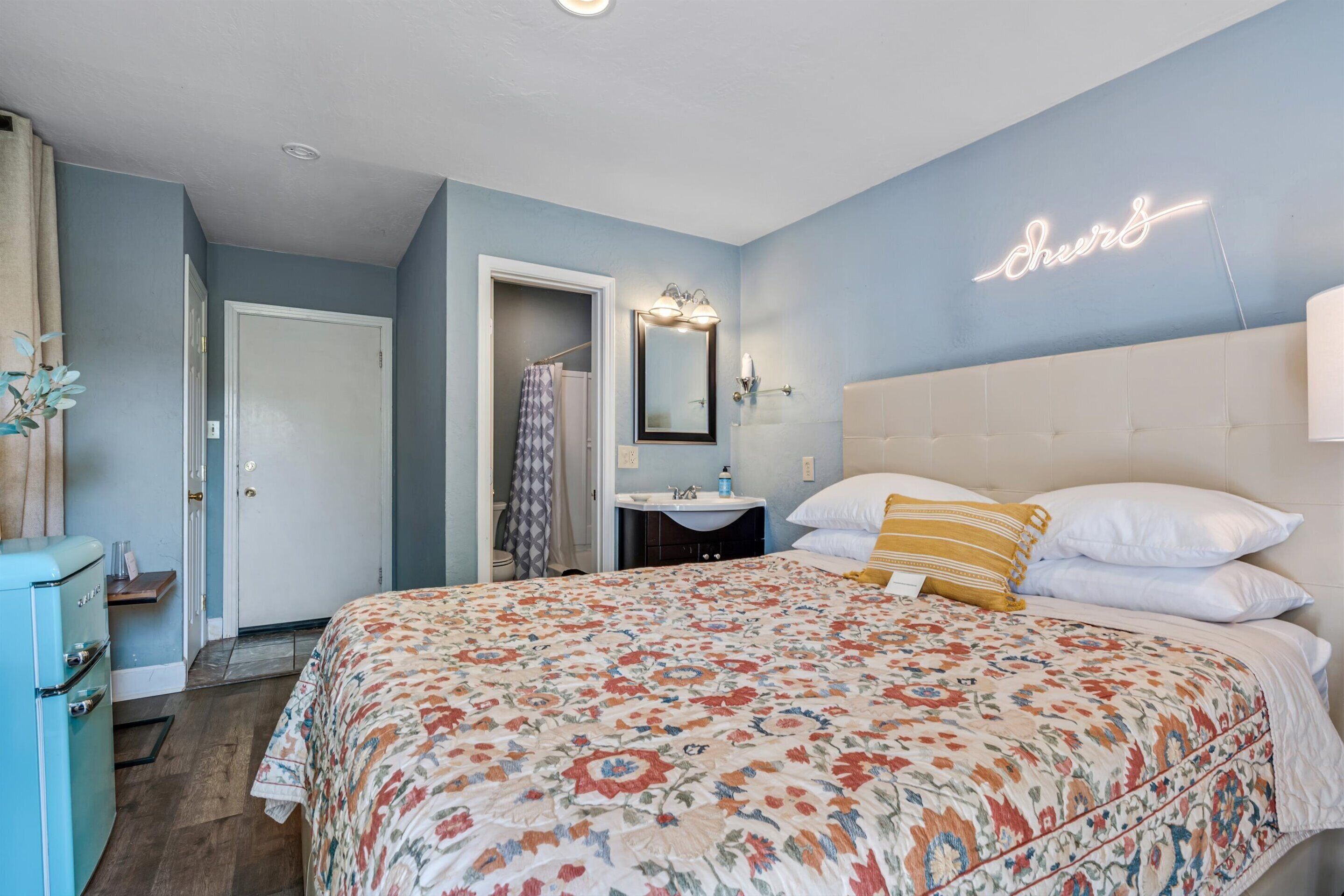 A bedroom with a floral bedspread, a neon sign on the wall, a white headboard, and a vintage-style dresser.