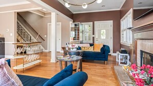 A spacious living room with a blue sofa, a coffee table, and a chandelier. A staircase is visible in the background leading to another room.