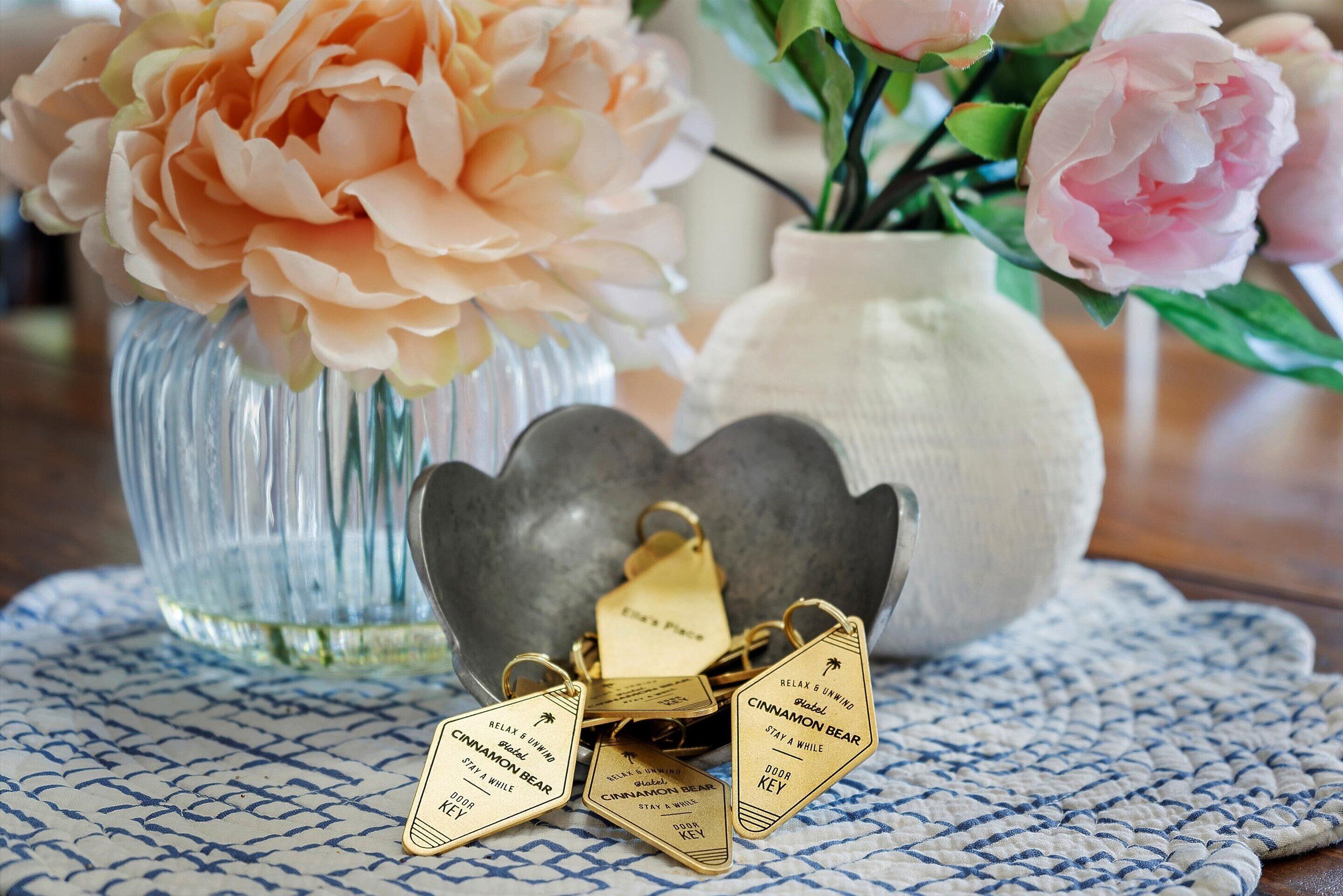 A table with a blue patterned tablecloth, a glass vase with peach flowers, a white vase with pink flowers, and a metal key holder with engraved keys.