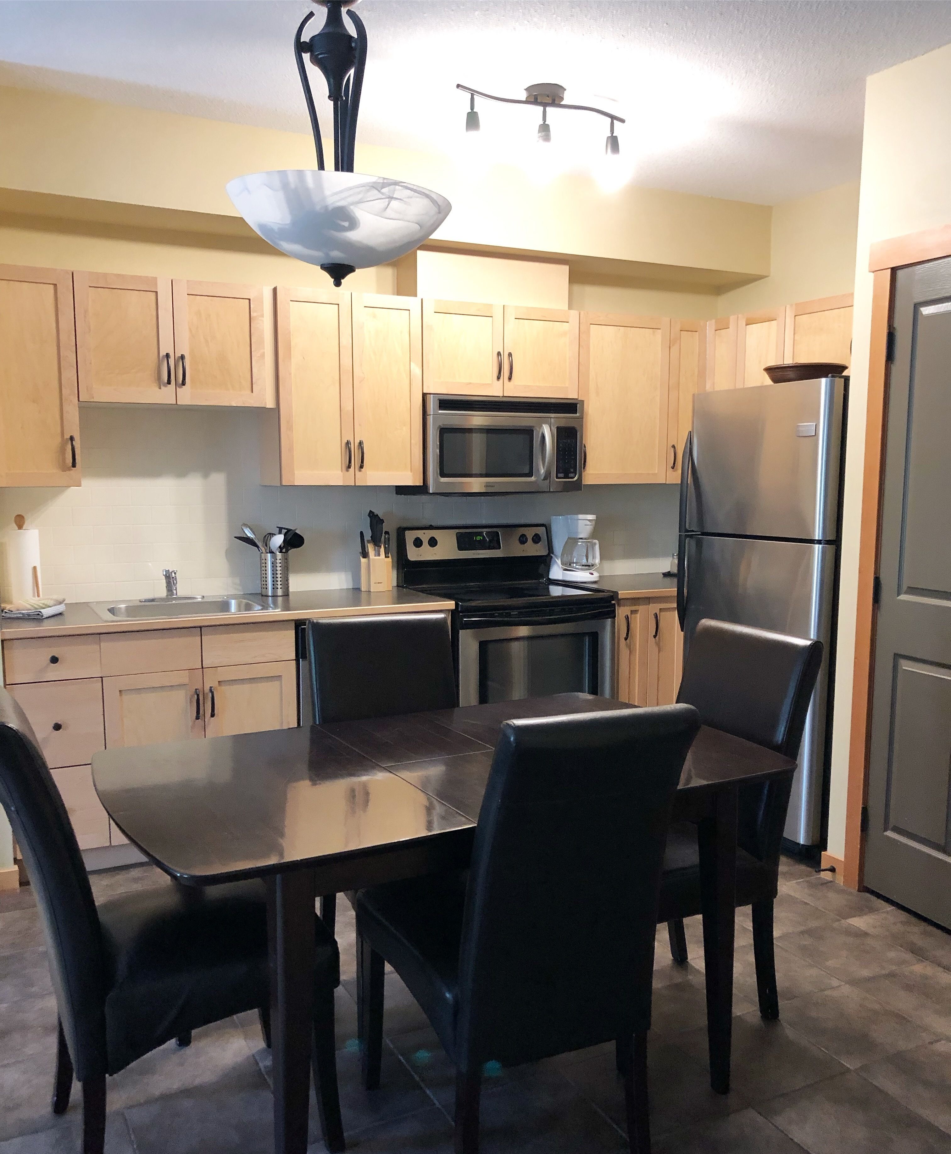 A modern kitchen with wooden cabinets, a stainless steel refrigerator, a microwave, an oven, a coffee maker, and a dining table with chairs.
