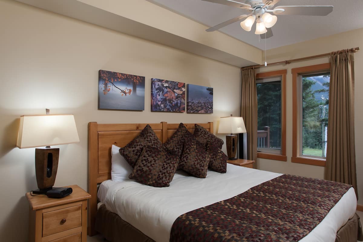 A neatly arranged bedroom with a bed, two bedside tables with lamps, a ceiling fan, and three framed artworks on the wall.