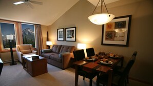 Habitación de hotel con zona de comedor, sofá, sillón, mesa para dos personas y cuadros en la pared.