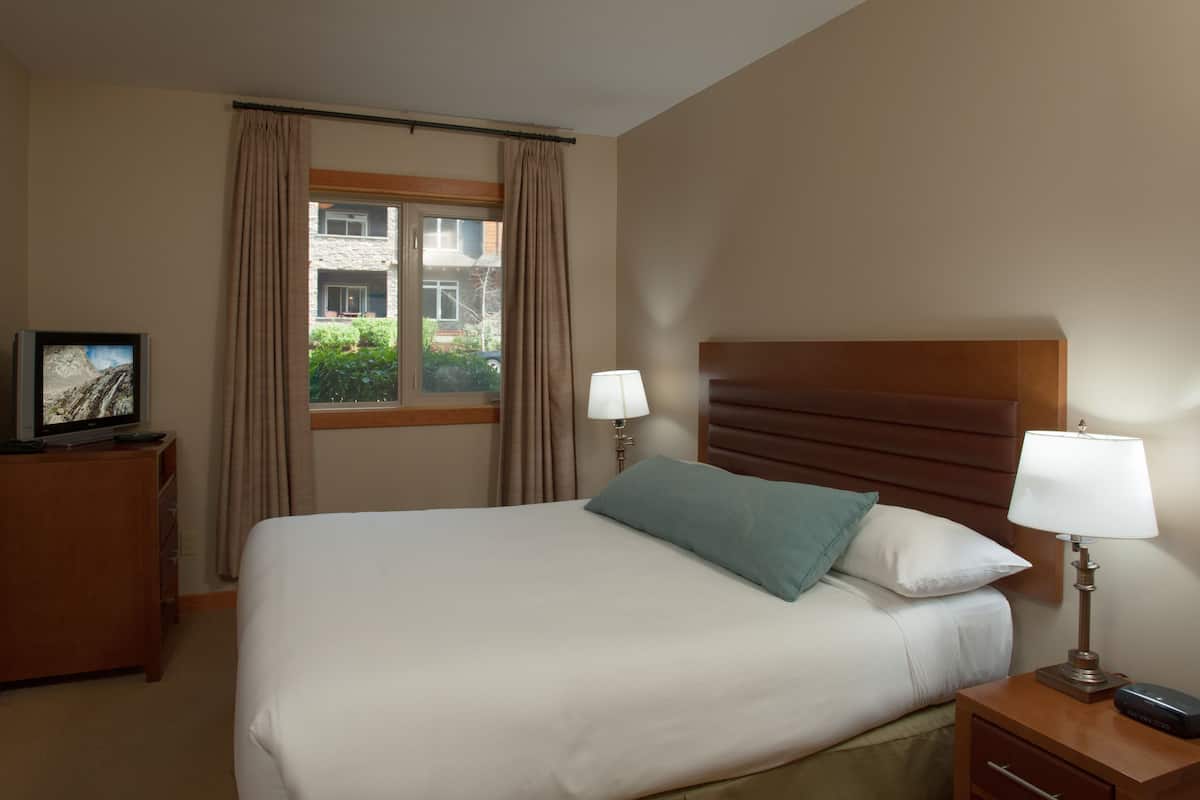 A hotel room with a bed, two bedside lamps, a TV on a wooden dresser, and a window with curtains.