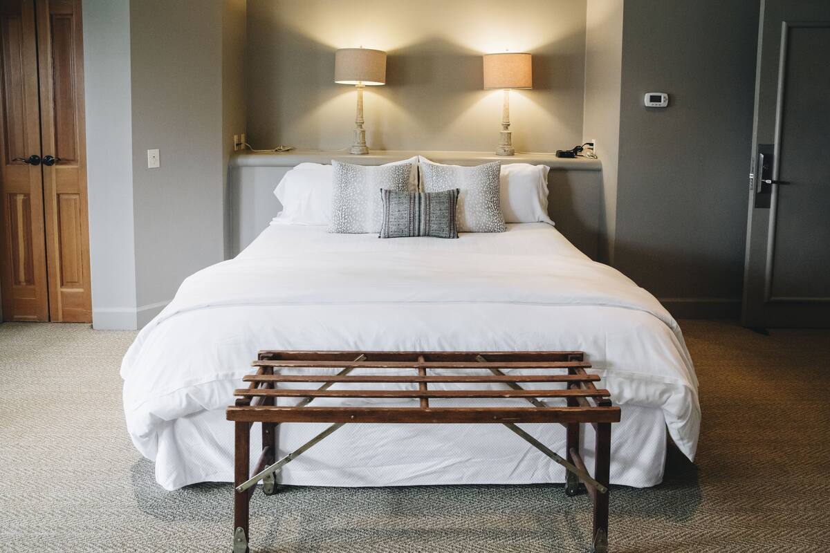 A neatly made bed with white linens and decorative pillows, a wooden bench in front, and two wall-mounted lamps.