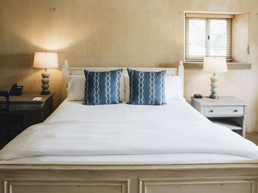 A neatly made bed with white linens and two blue patterned pillows, a bedside lamp, a small table with a lamp, and a window with blinds.