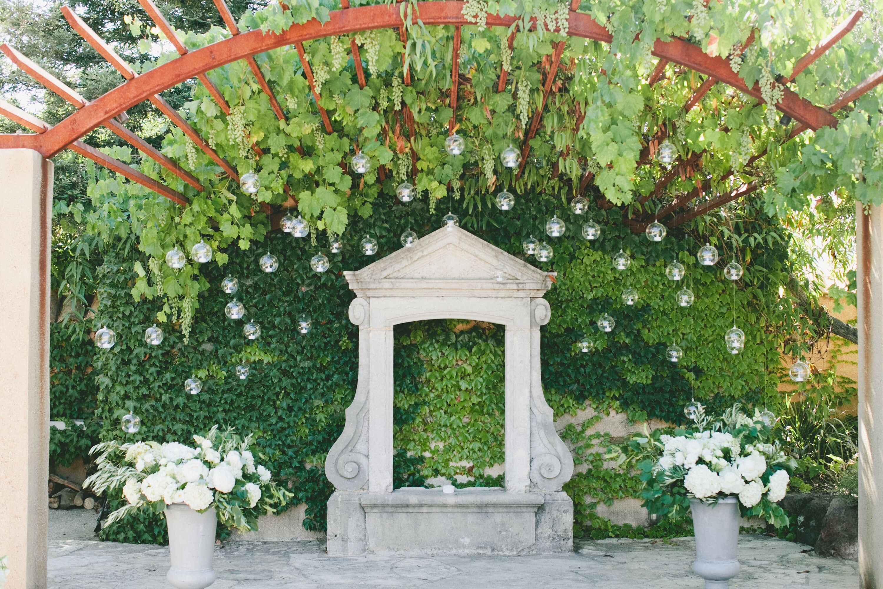 A stone archway with a white flower arrangement on either side, surrounded by greenery and hanging glass orbs.