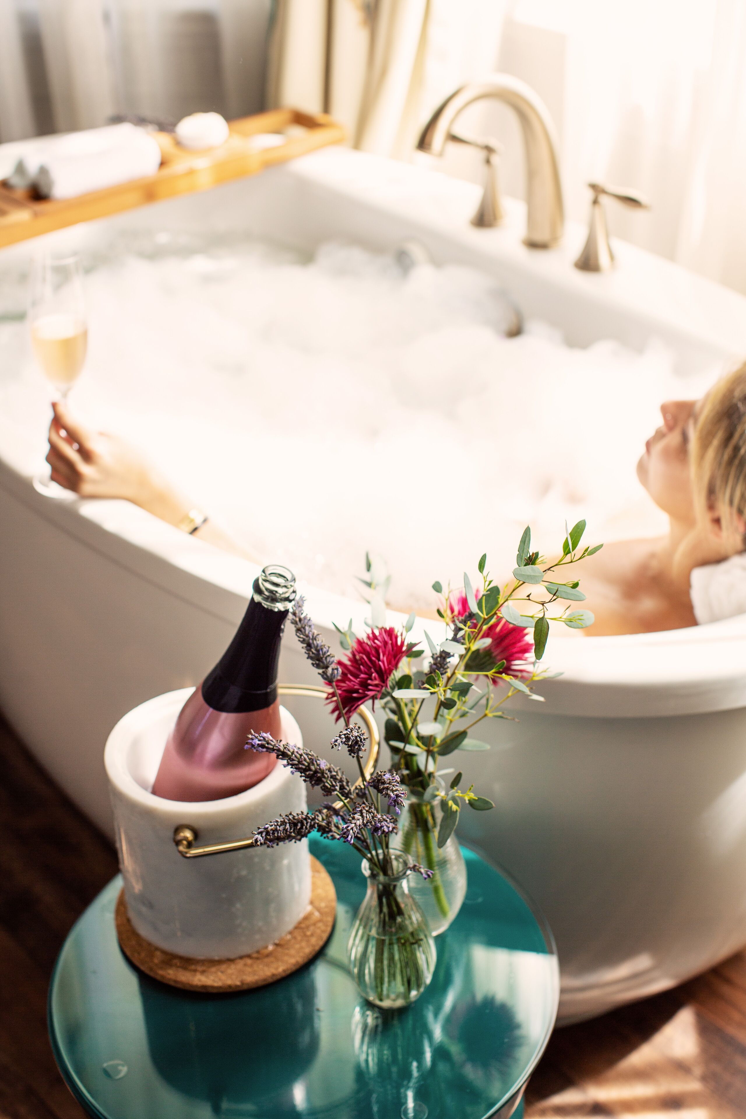 A bathtub with a person sitting in it, a bottle of champagne, and a vase with flowers on a table beside the tub.