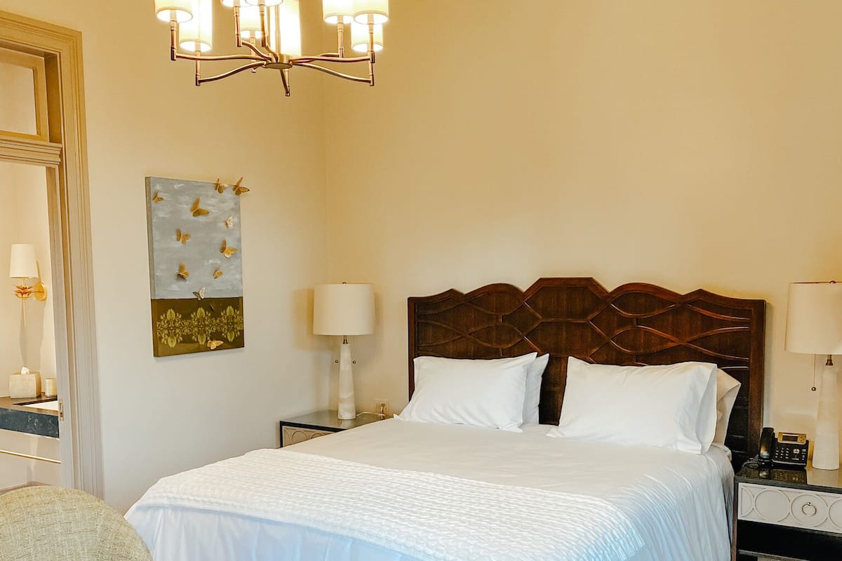 A hotel room with a large bed, two armchairs, a small table, a chandelier, and a painting on the wall.