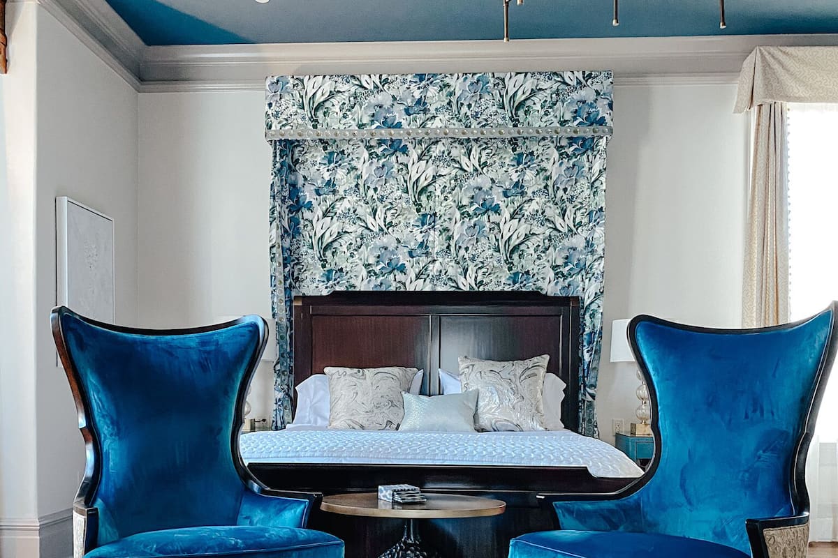 A bedroom with a large bed, two blue armchairs, a round table, and a patterned wallpaper.