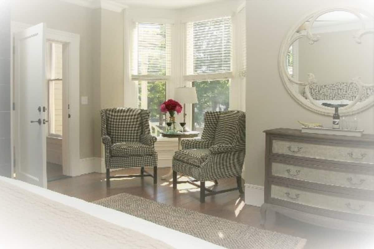 A room with two checkered armchairs, a small table with a vase of flowers, a dresser with a mirror, and a window with blinds.