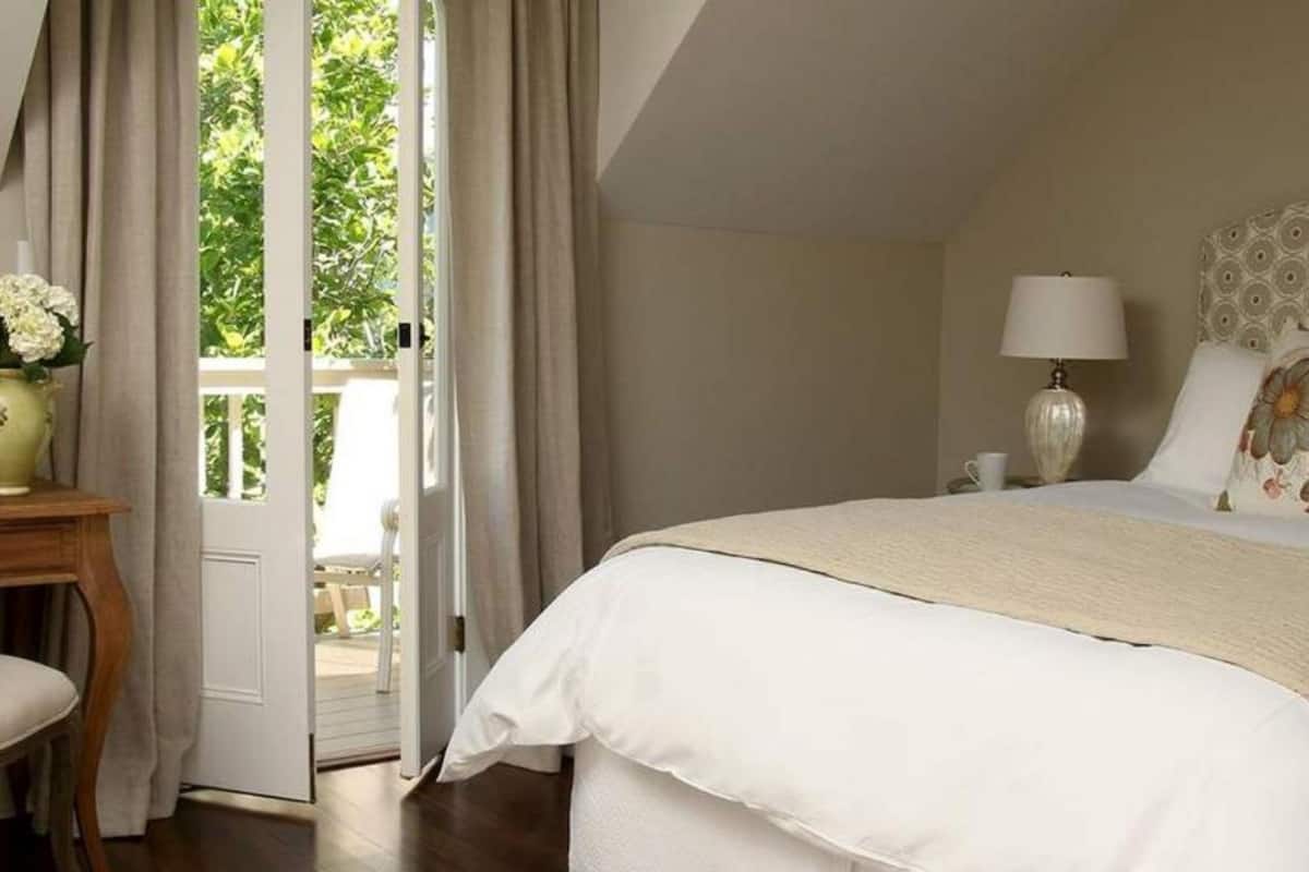A bedroom with a large bed, a nightstand, a lamp, and a view of greenery through an open door.
