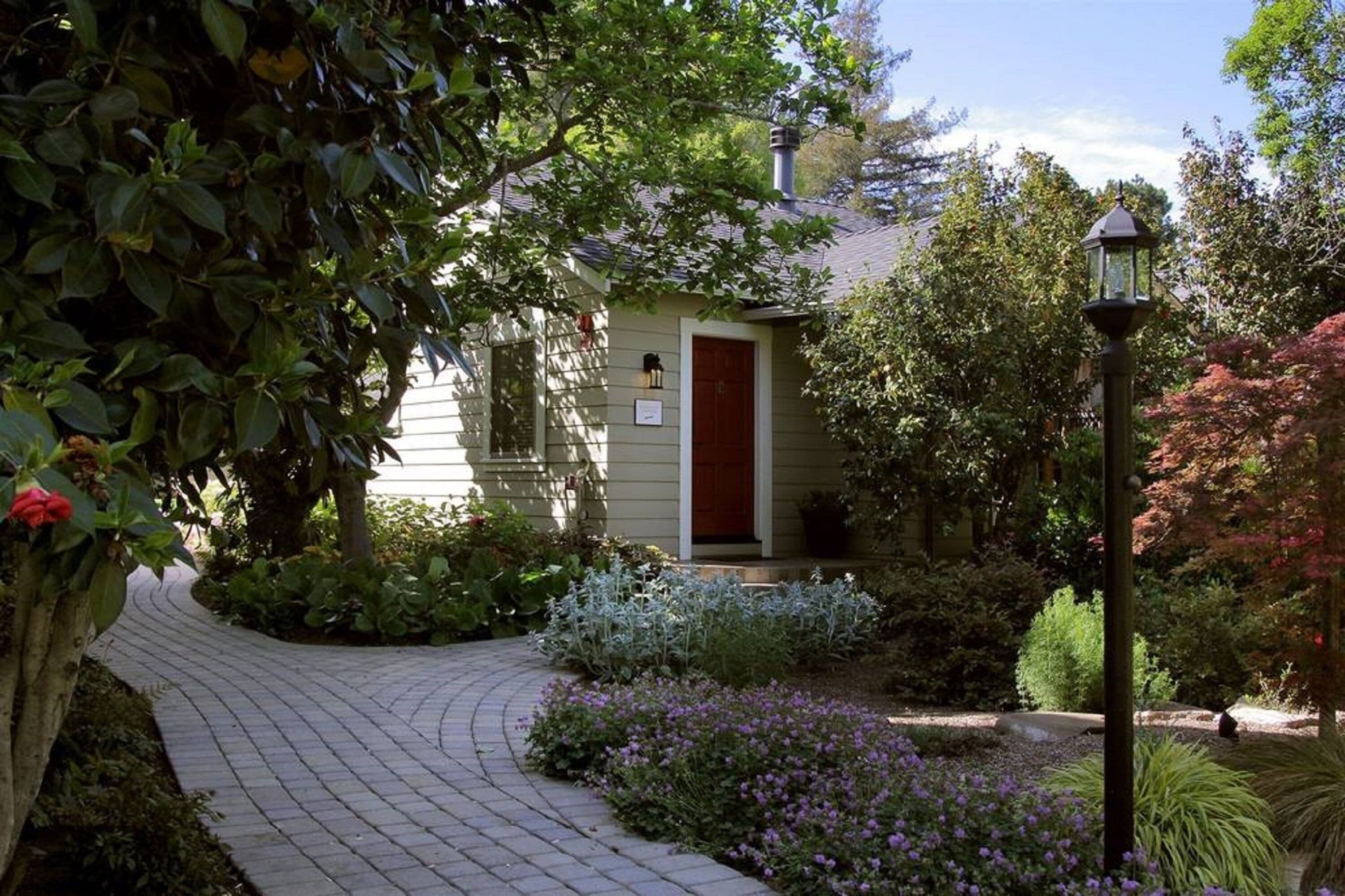 A house with a red door, surrounded by a garden with various plants and a paved pathway.