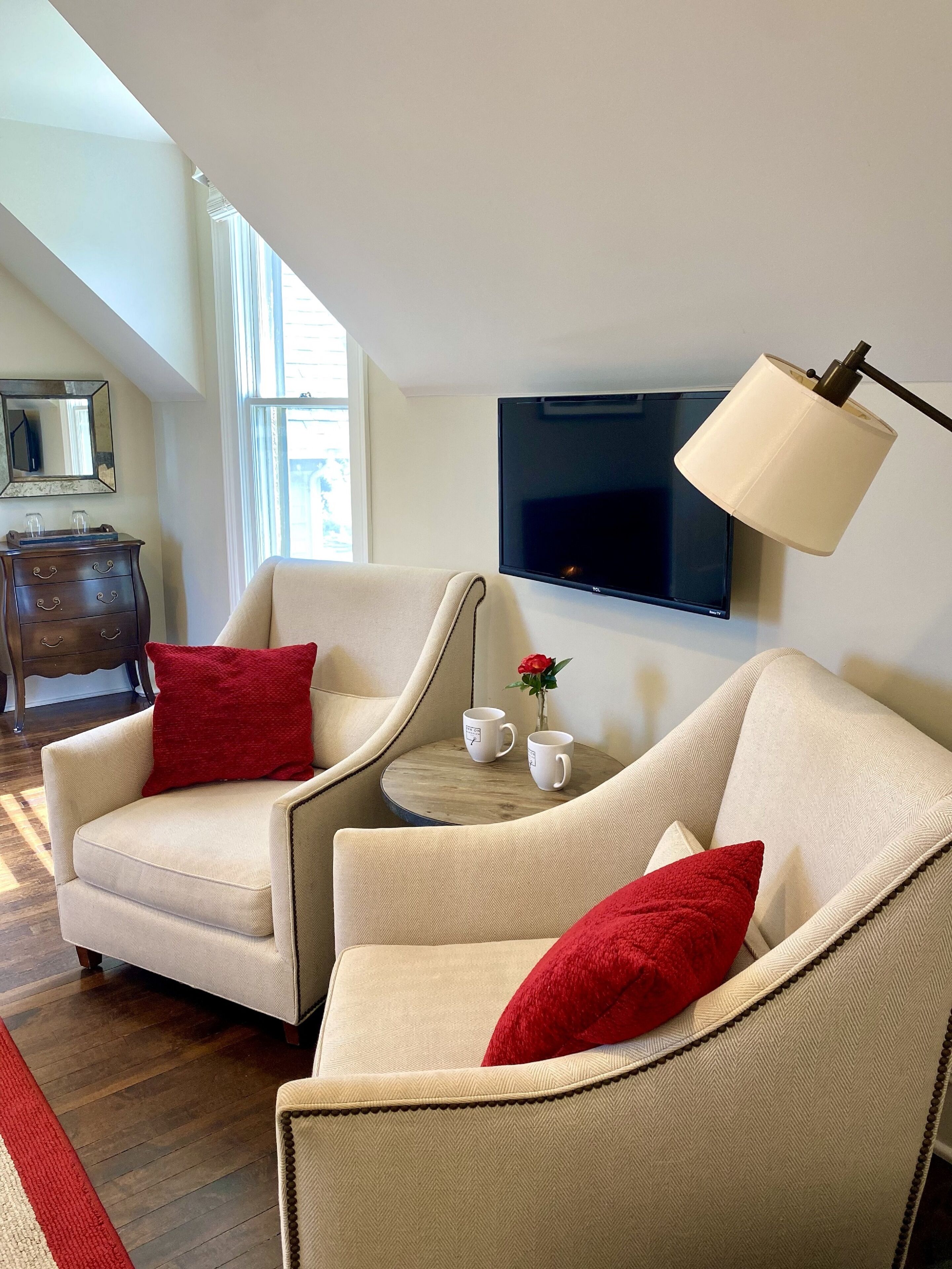 Un salón con dos sillones beige, una mesita redonda con dos tazas blancas, una rosa roja y un televisor de pantalla plana montado en la pared.