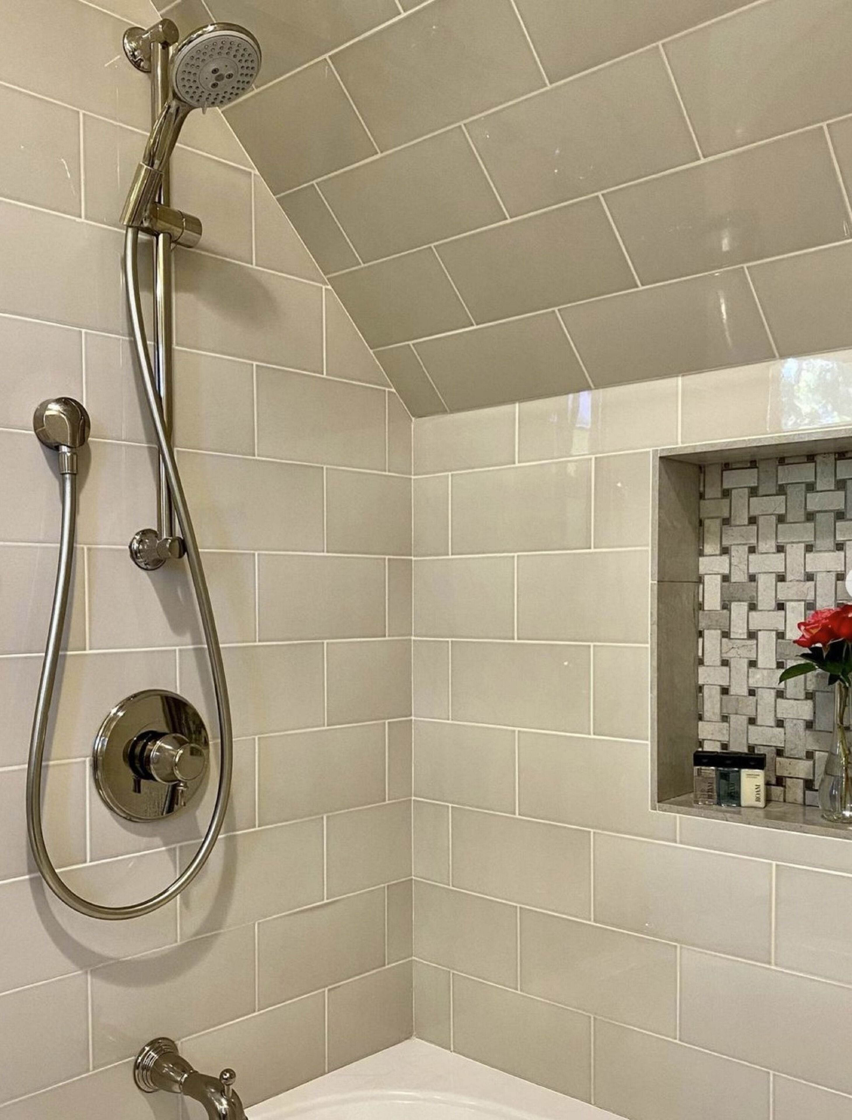 A bathroom with white tiled walls, a shower with a handheld showerhead, a wall-mounted faucet, and a small shelf with toiletries.