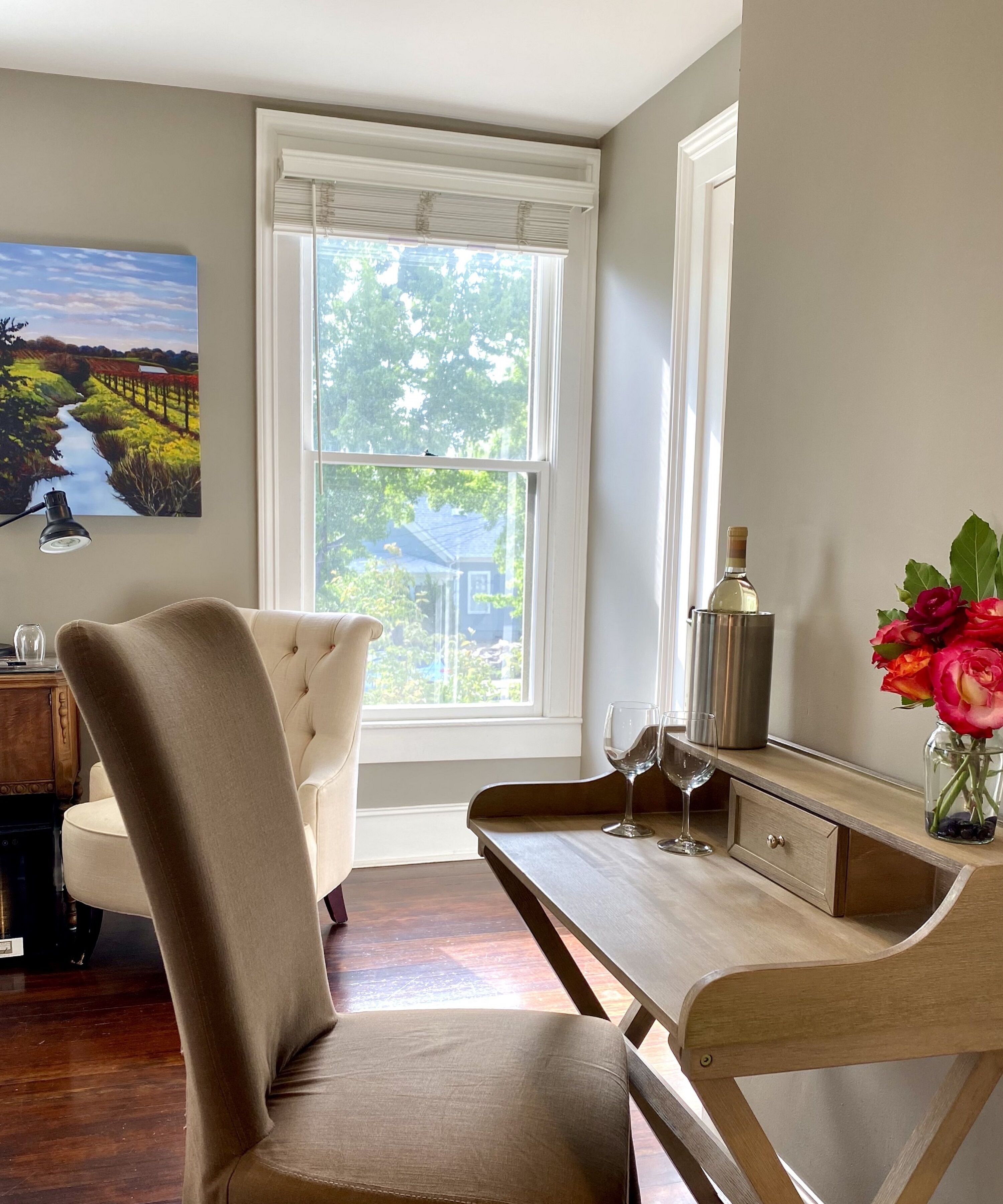 Une pièce avec un bureau en bois, une chaise rembourrée, un tableau, une bouteille de vin et un vase rempli de fleurs.
