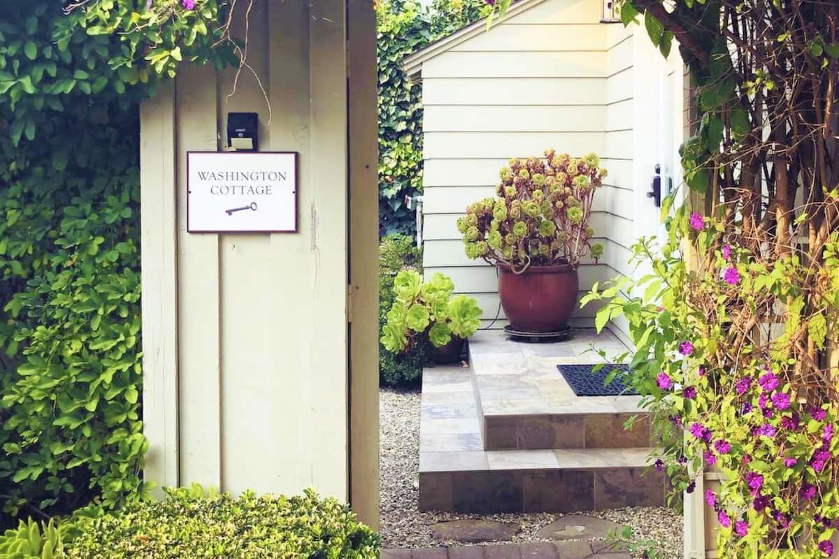 A garden entrance with a sign reading "Washington Cottage," a potted plant, and a pathway leading to a white building.