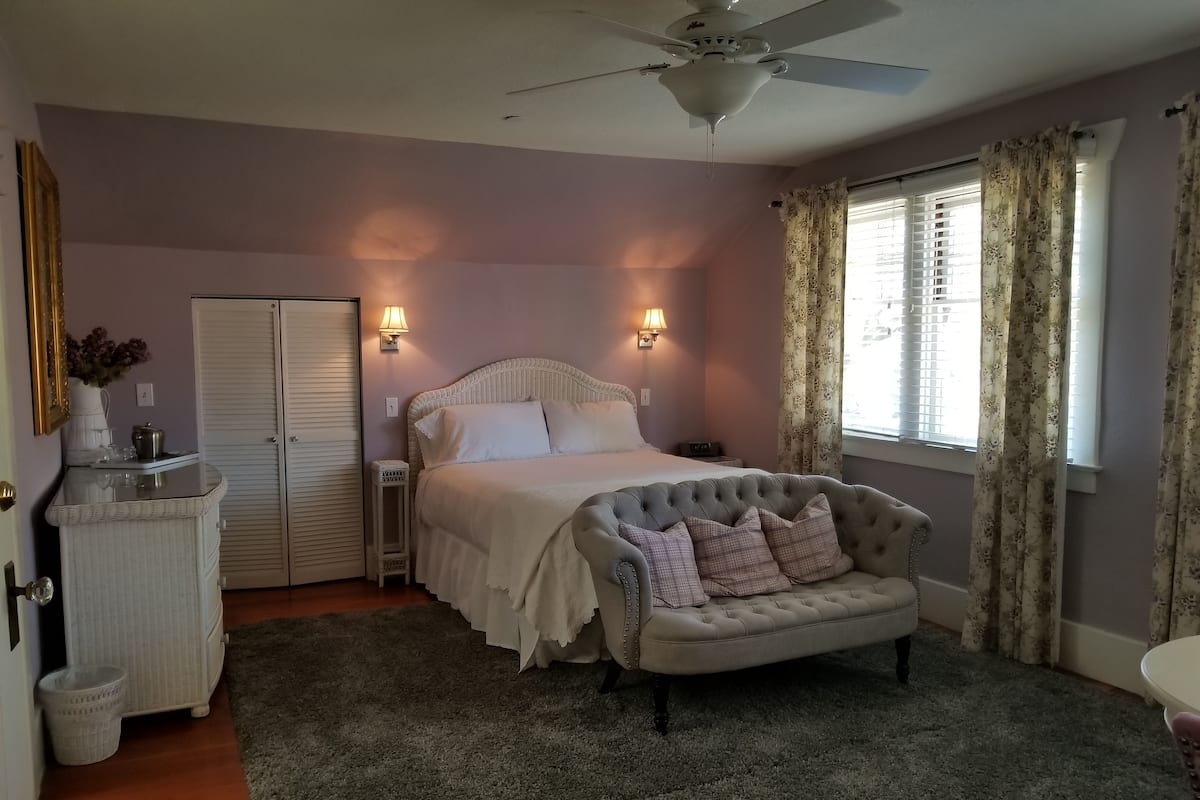 A bedroom with a bed, a dresser, a vanity, a ceiling fan, and a window with curtains.