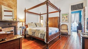 Una cama con dosel de madera y colcha, un escritorio de madera con lámpara, una silla y un armario de madera en una habitación con piso de madera y pared de ladrillo.