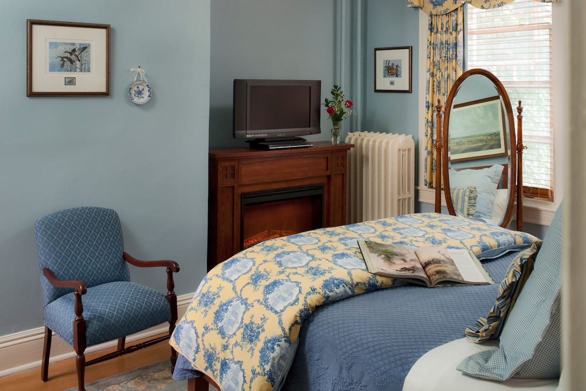 A bedroom with a bed, a chair, a television on a wooden stand, a fireplace, and a window with curtains.