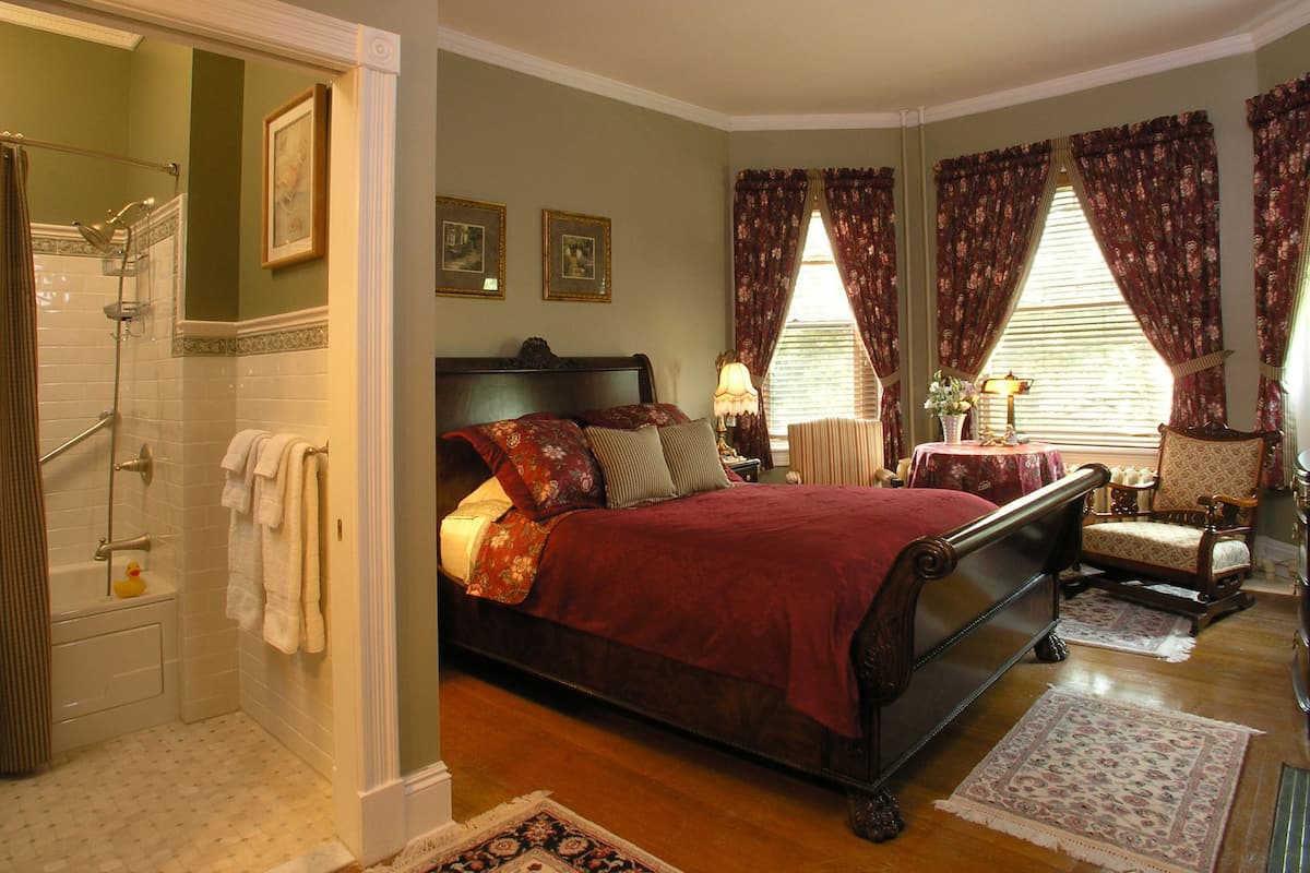 A bedroom with a large bed, a sitting area with a chair, a small table, and a shower area.