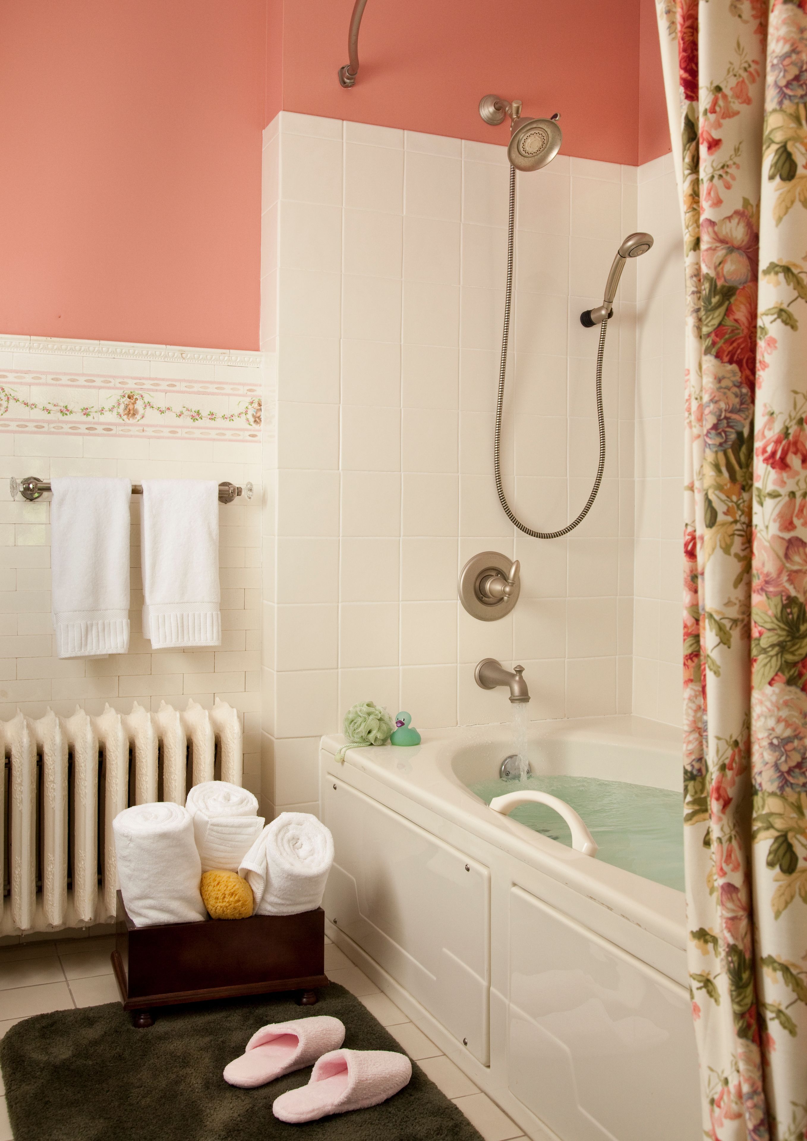A bathroom with a bathtub, shower, and a radiator. There are towels and slippers on the floor.