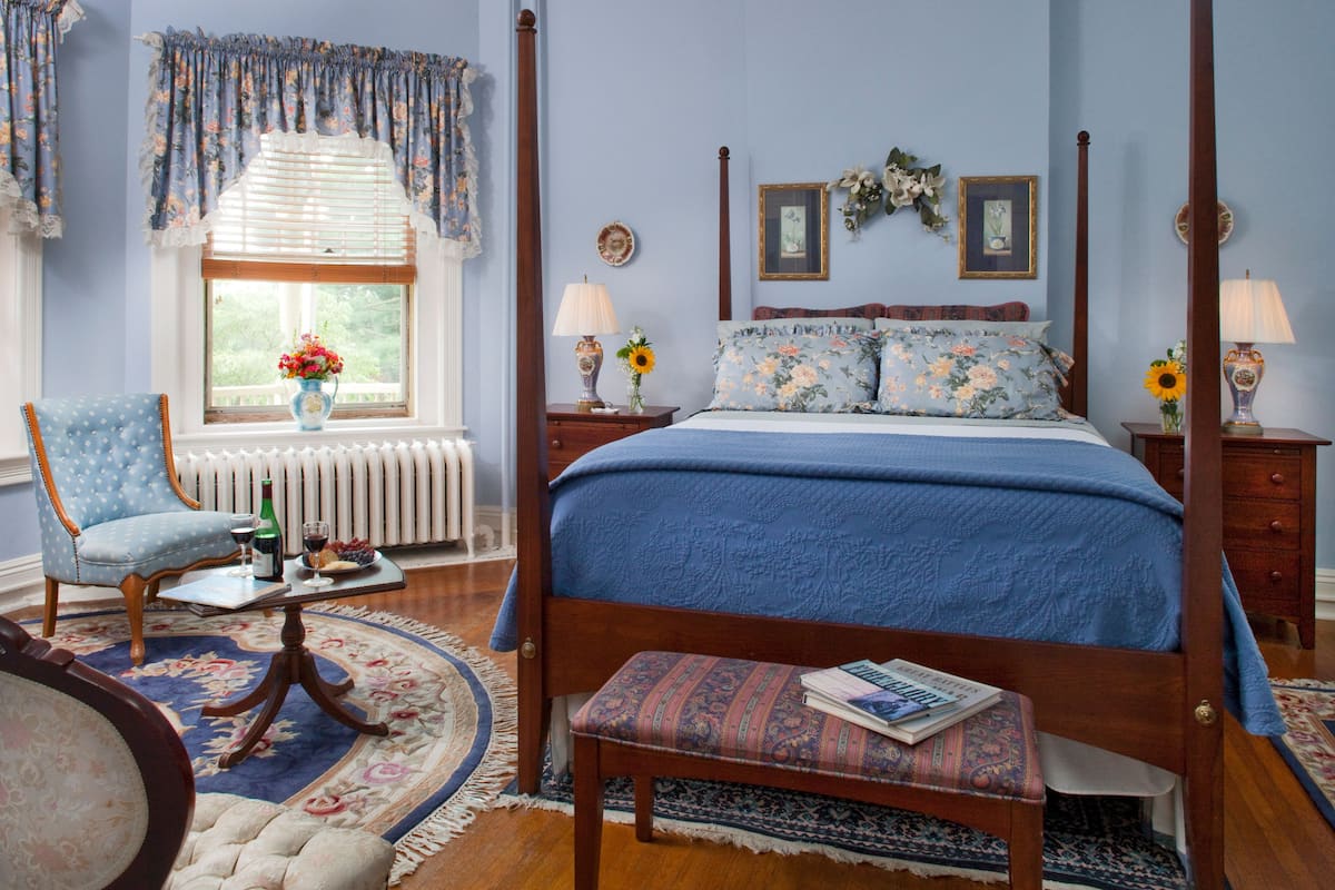 A bedroom with a four-poster bed, a wooden bench, a chair, a bedside table with a lamp, and a window with curtains.