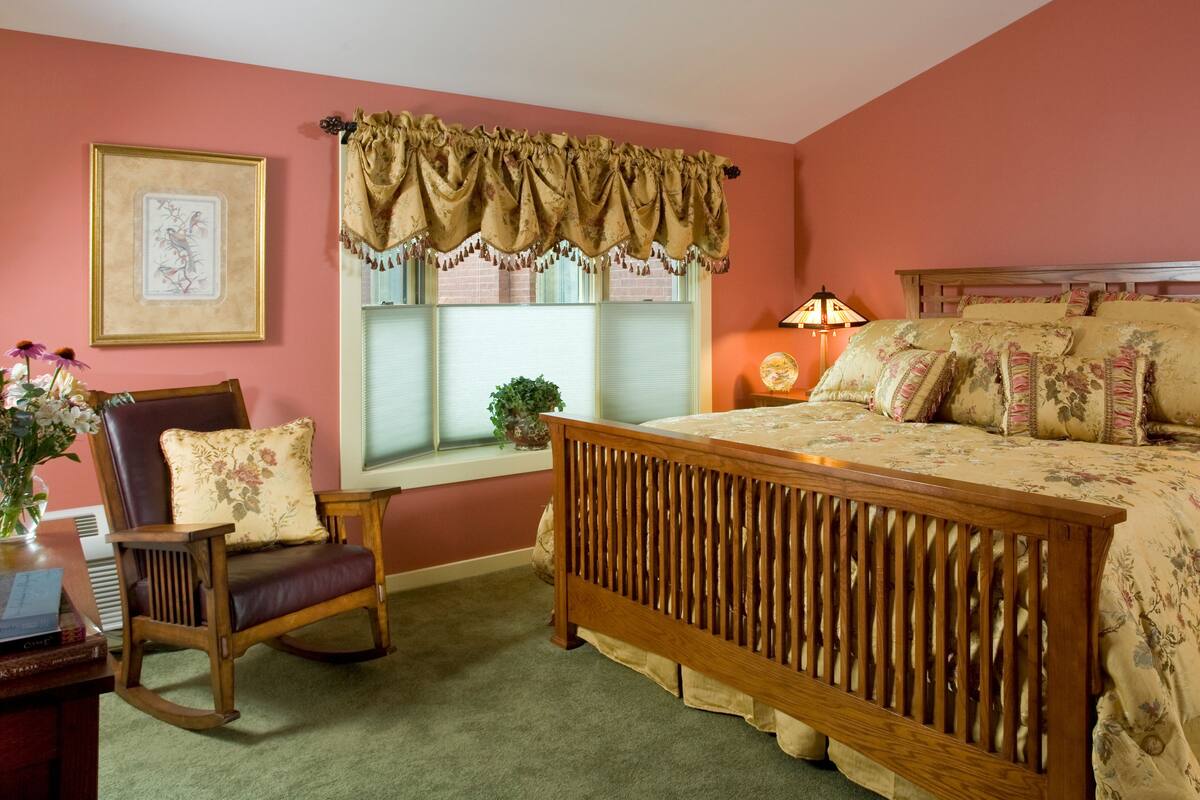 A bedroom with a wooden bed, a rocking chair, a bedside table with a lamp, and a window with curtains.