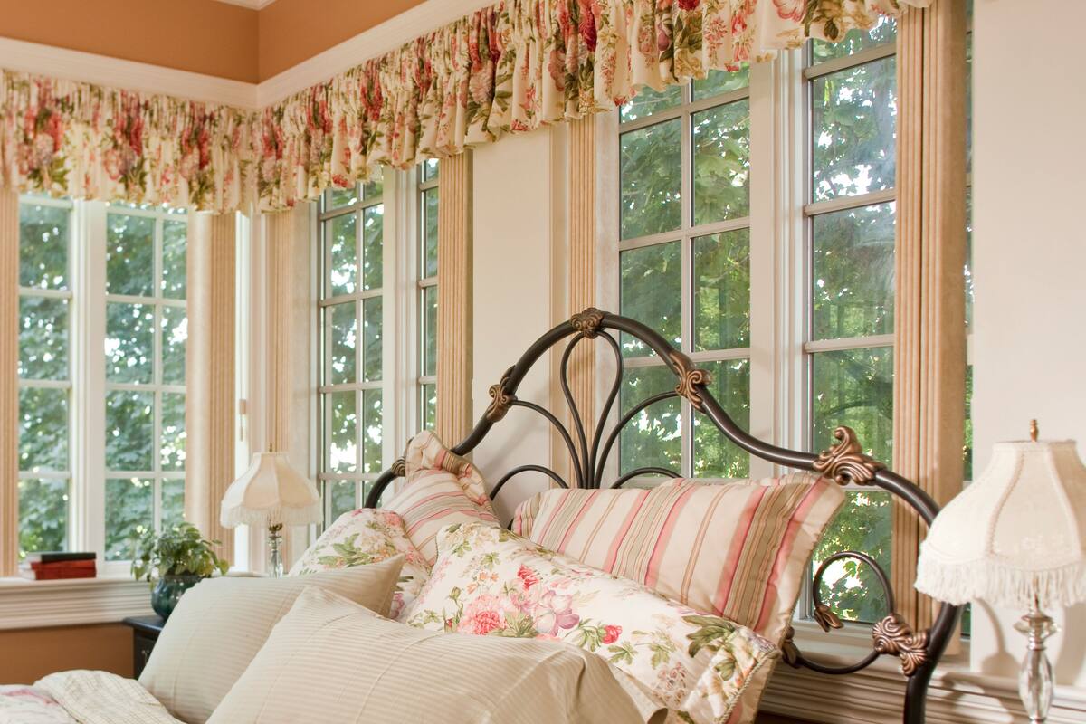 A bedroom with a bed, floral bedding, a nightstand with a lamp, and a dresser with books.