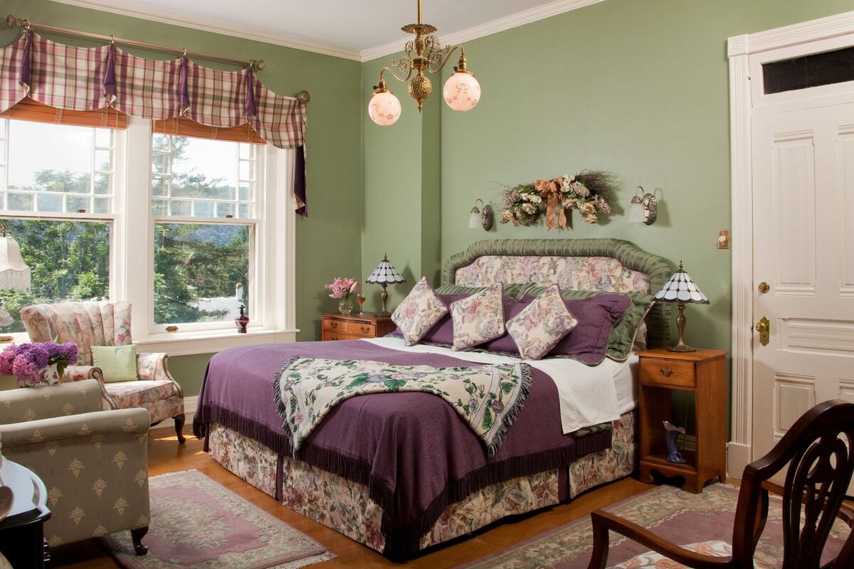 A bedroom with a large bed, floral-patterned bedding, a view of greenery through the window, and a vintage chair.