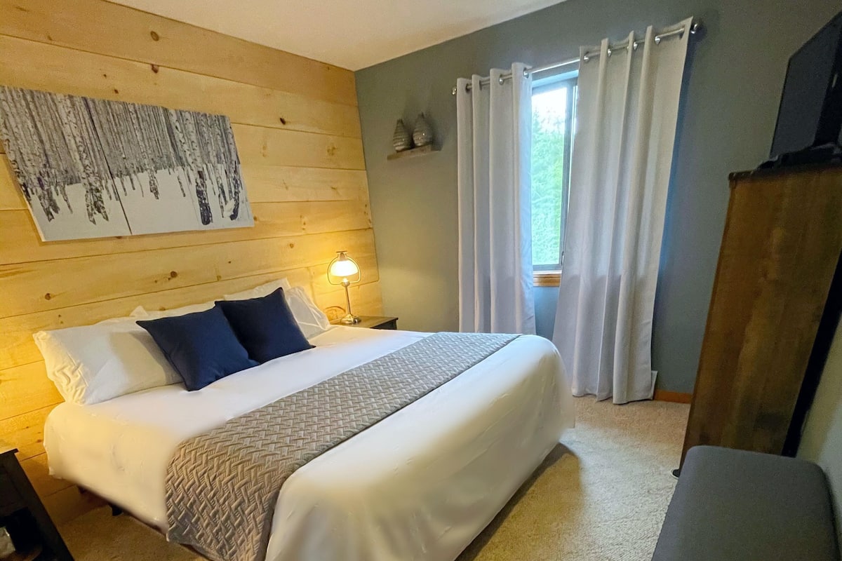 A bedroom with a wooden headboard, a bed with white bedding and blue pillows, a bedside lamp, a window with curtains, and a wooden wardrobe.