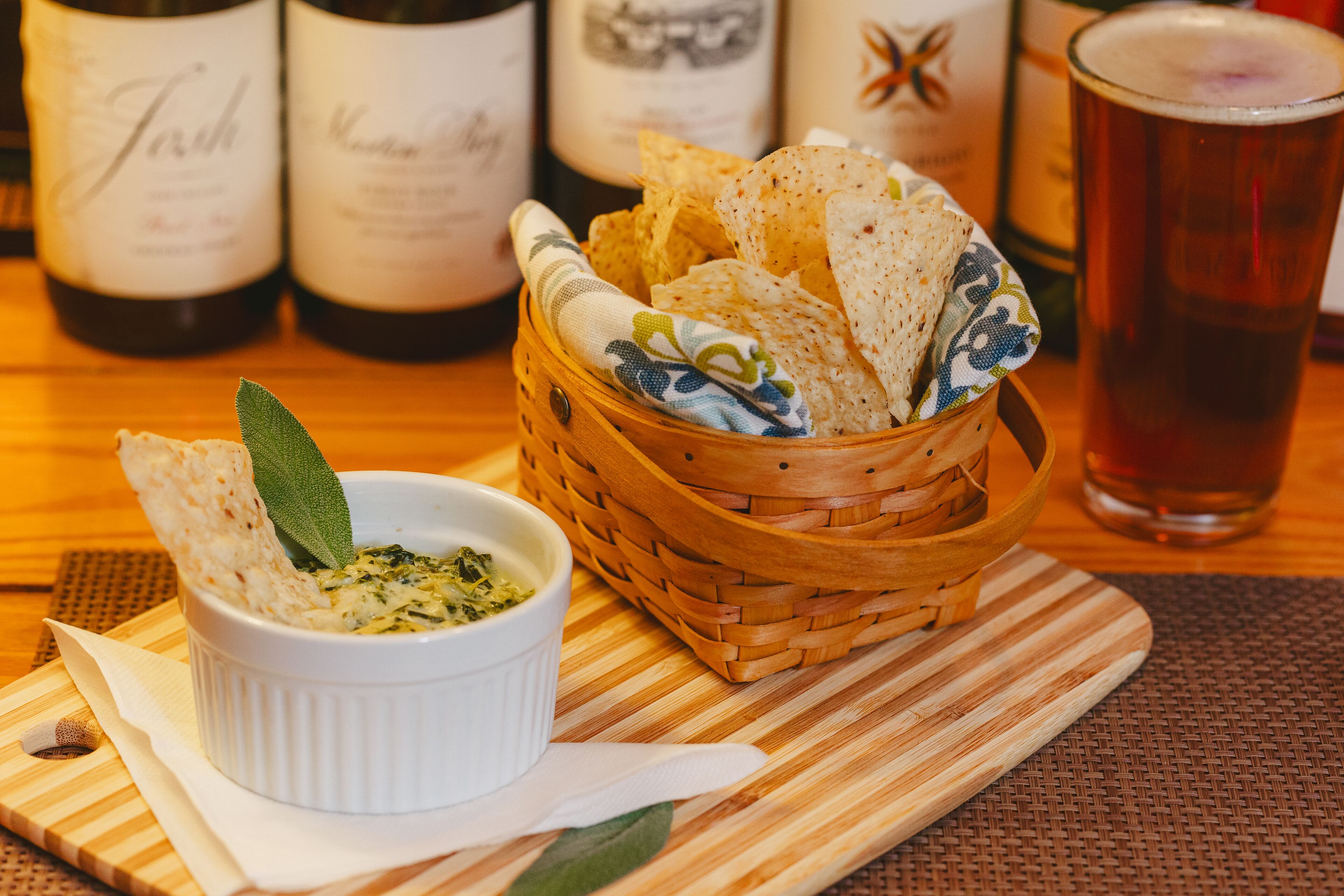 A wooden table with a white bowl of dip, a basket of flatbread, and a glass of beer.