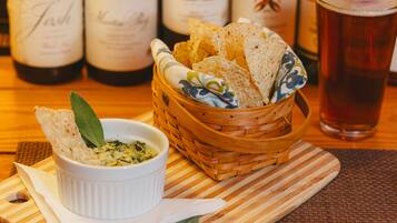 A wooden table with a white bowl of dip, a basket of flatbread, and a glass of beer.