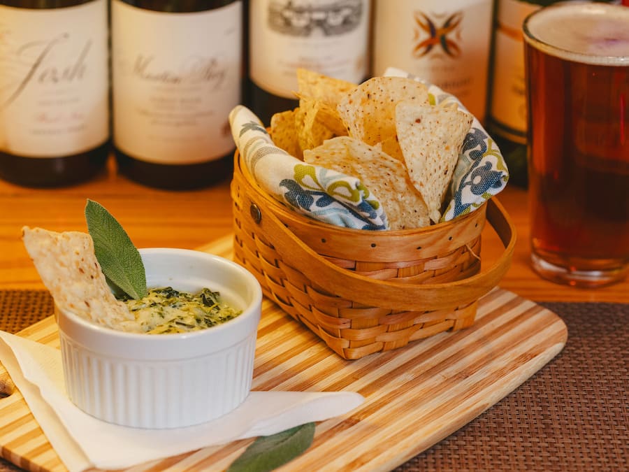 A wooden table with a white bowl of dip, a basket of flatbread, and a glass of beer.