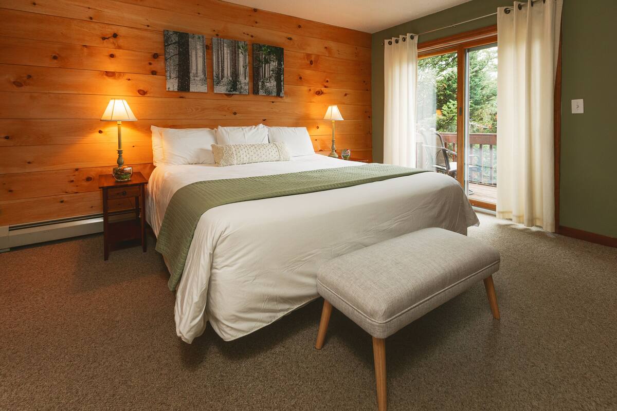 Un dormitorio con cabecera de madera, una cama grande con ropa blanca, dos lámparas de noche y vistas al exterior a través de una puerta corrediza de vidrio.