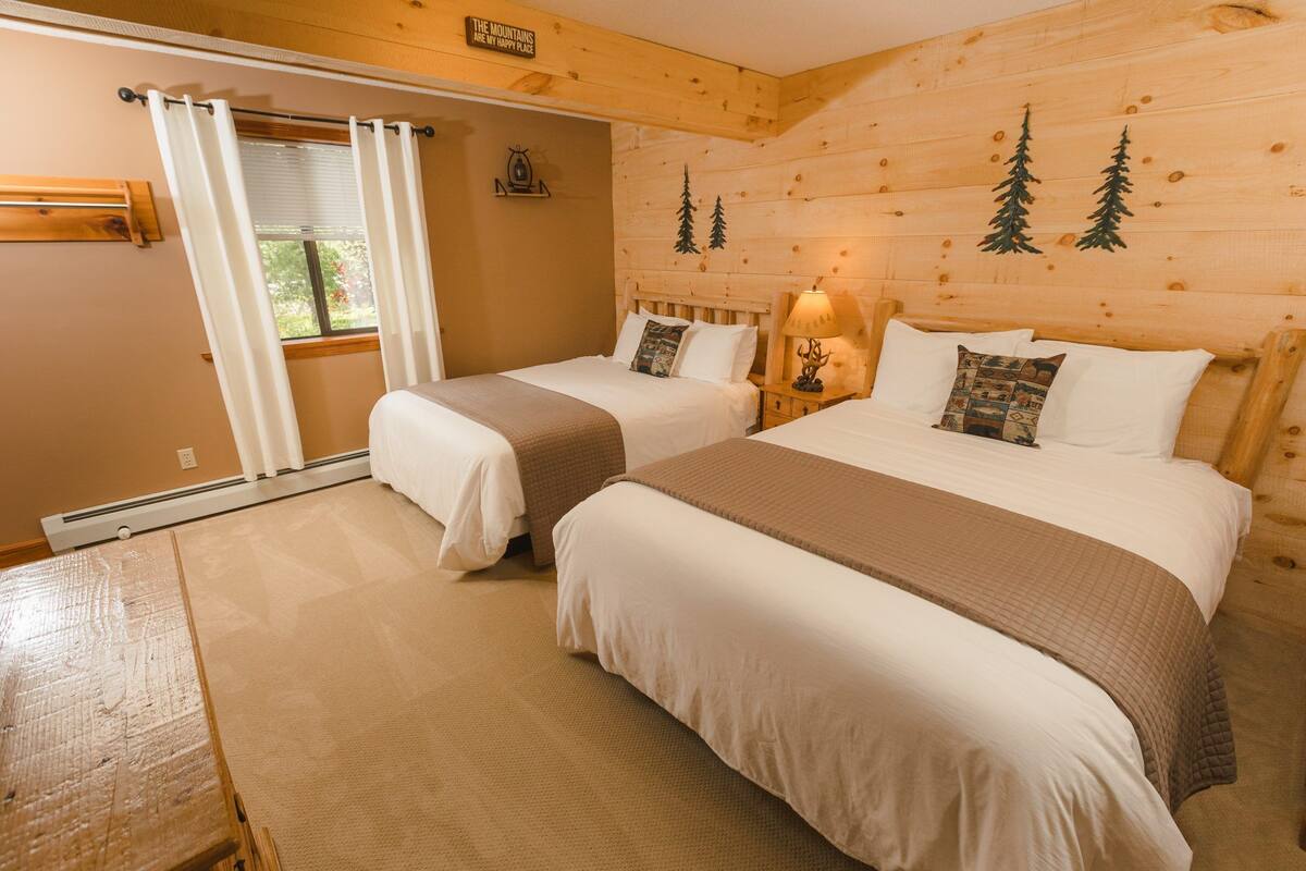 A cozy cabin-style room with two beds, a wooden wall with tree decals, a window with curtains, and a small table.