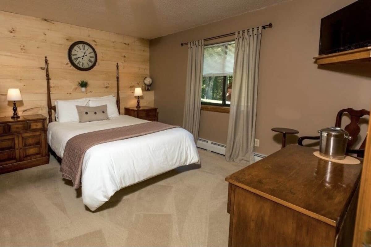 A bedroom with a wooden headboard, a bed with white bedding, two bedside tables with lamps, a clock, a window with curtains, and a small table with a kettle.