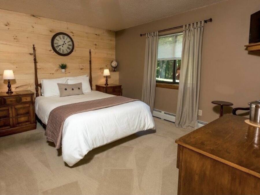 Une chambre à coucher avec une tête de lit en bois, un lit avec une literie blanche, deux tables de chevet avec des lampes, une horloge, une fenêtre avec des rideaux et une petite table avec une bouilloire.