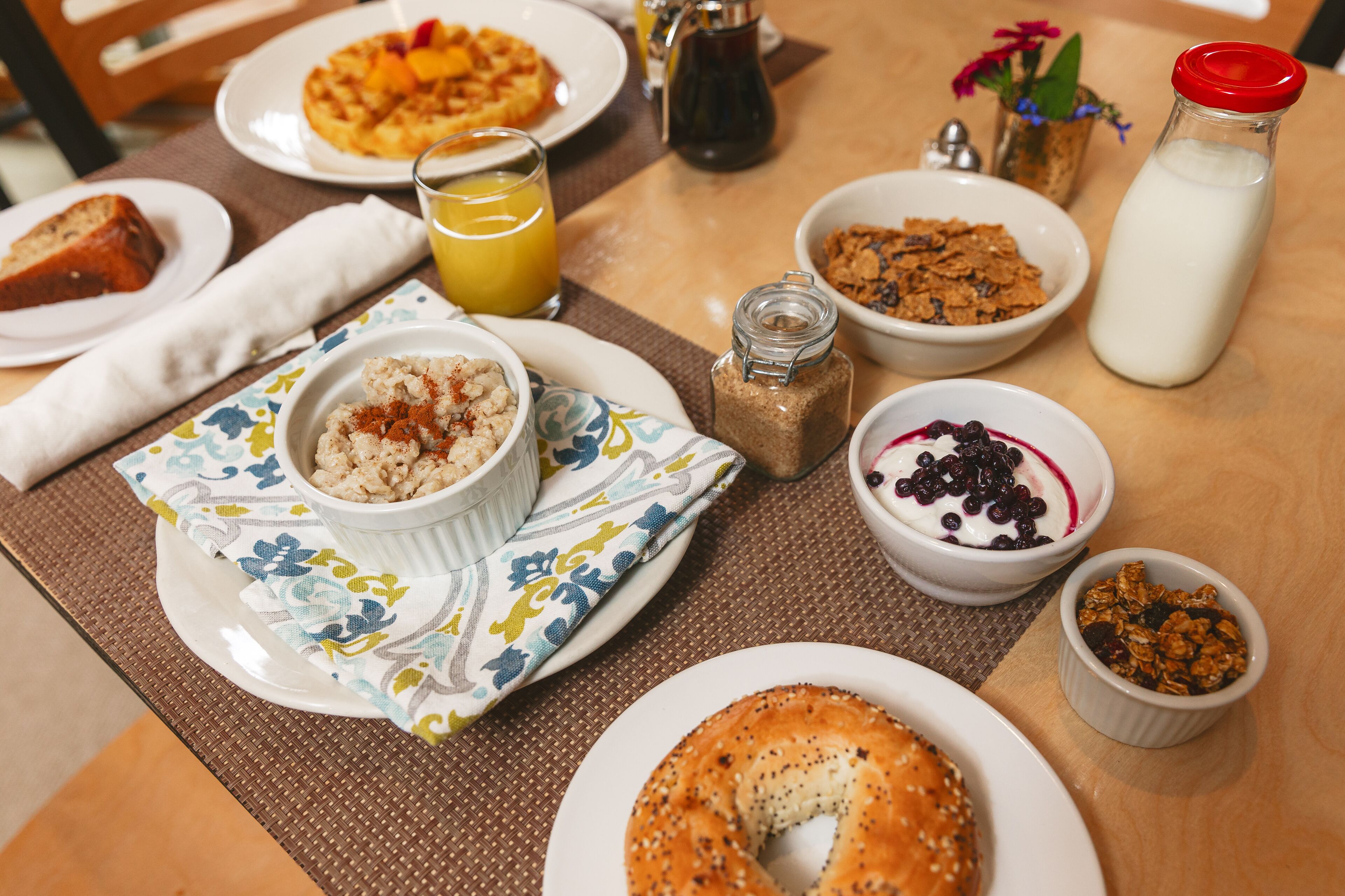 A breakfast table with various food items including a bagel, cereal, and a glass of orange juice.