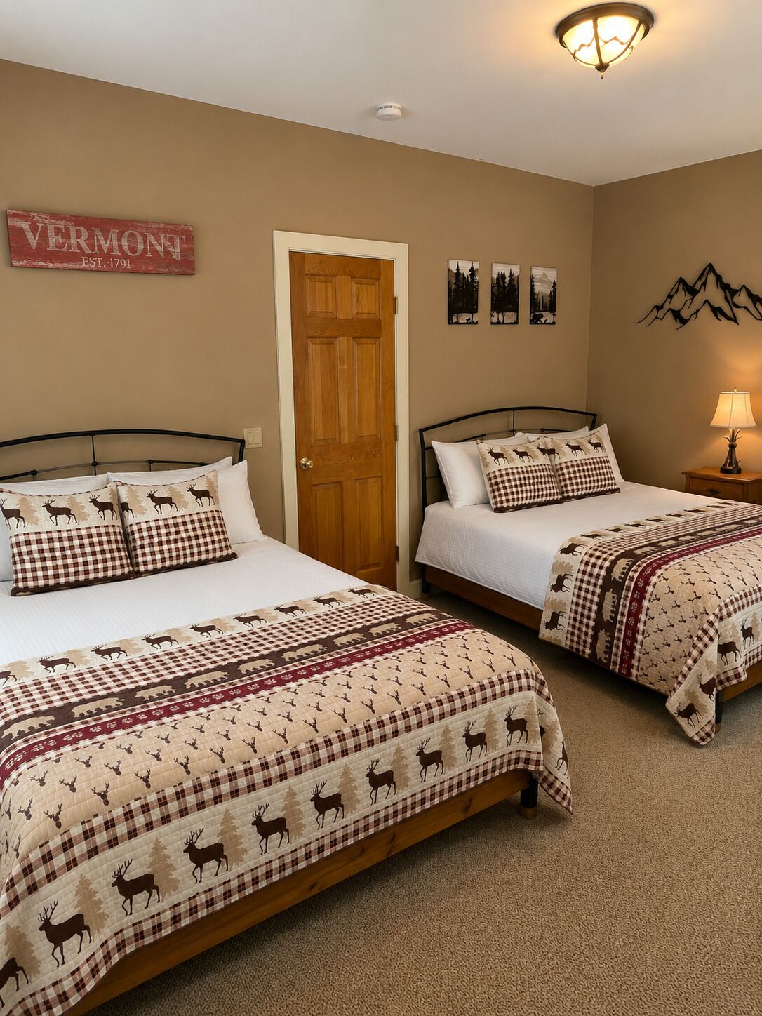 Habitación de hotel con dos camas, cada una con una colcha a cuadros y un cojín con estampado de ciervo. Se ve una puerta de madera, y hay decoraciones en las paredes, incluyendo un cartel que dice 'VERMONT EST. 1791' y una silueta de montaña.