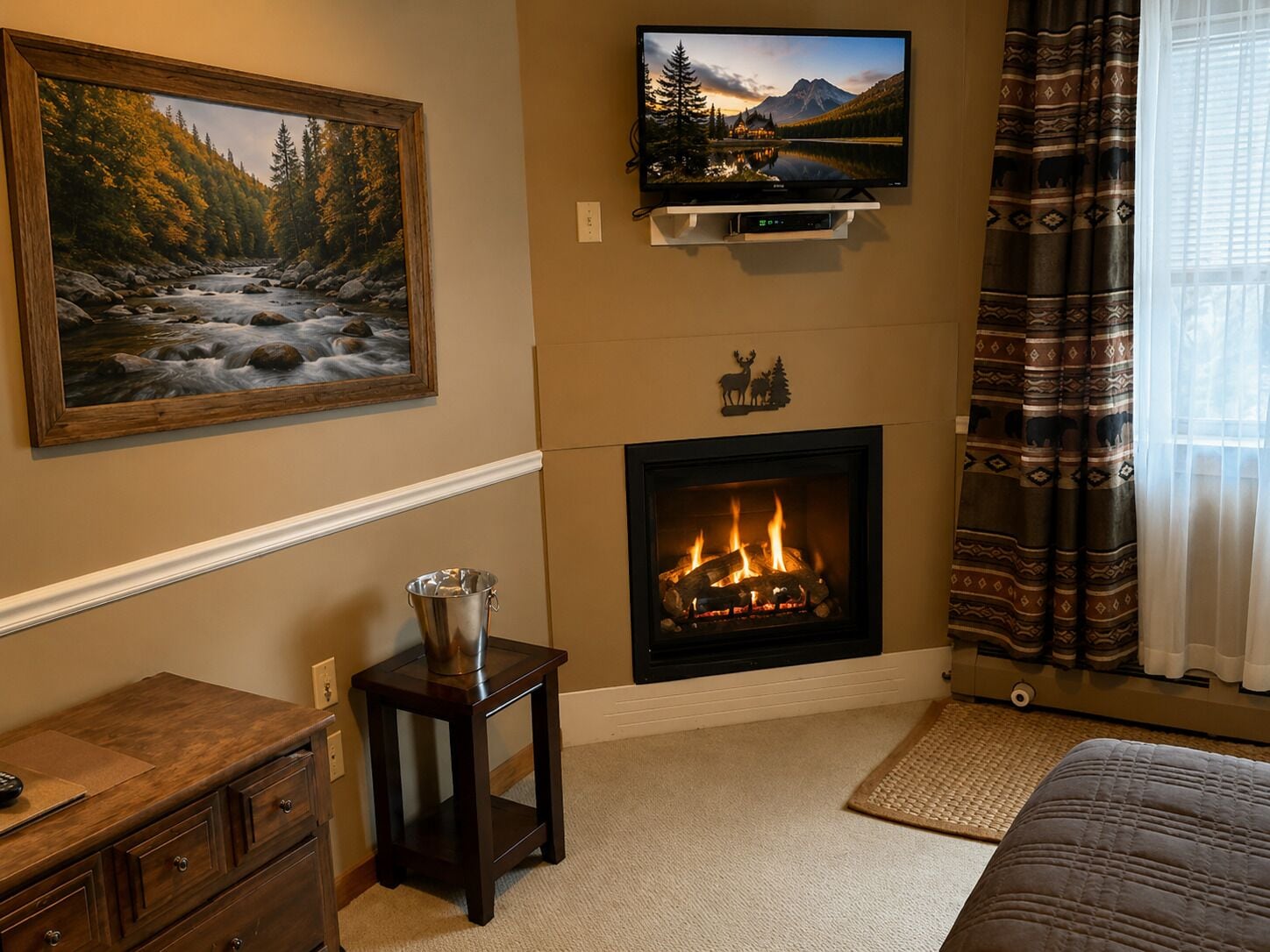 Una habitación con chimenea, un televisor montado encima, un cuadro enmarcado con escena de bosque y una cama con colcha estampada.