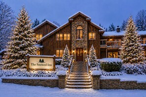 Un gran lodge de piedra y madera con un letrero que dice 'The Snowed Inn', rodeado de árboles cubiertos de nieve y un camino de entrada nevado.