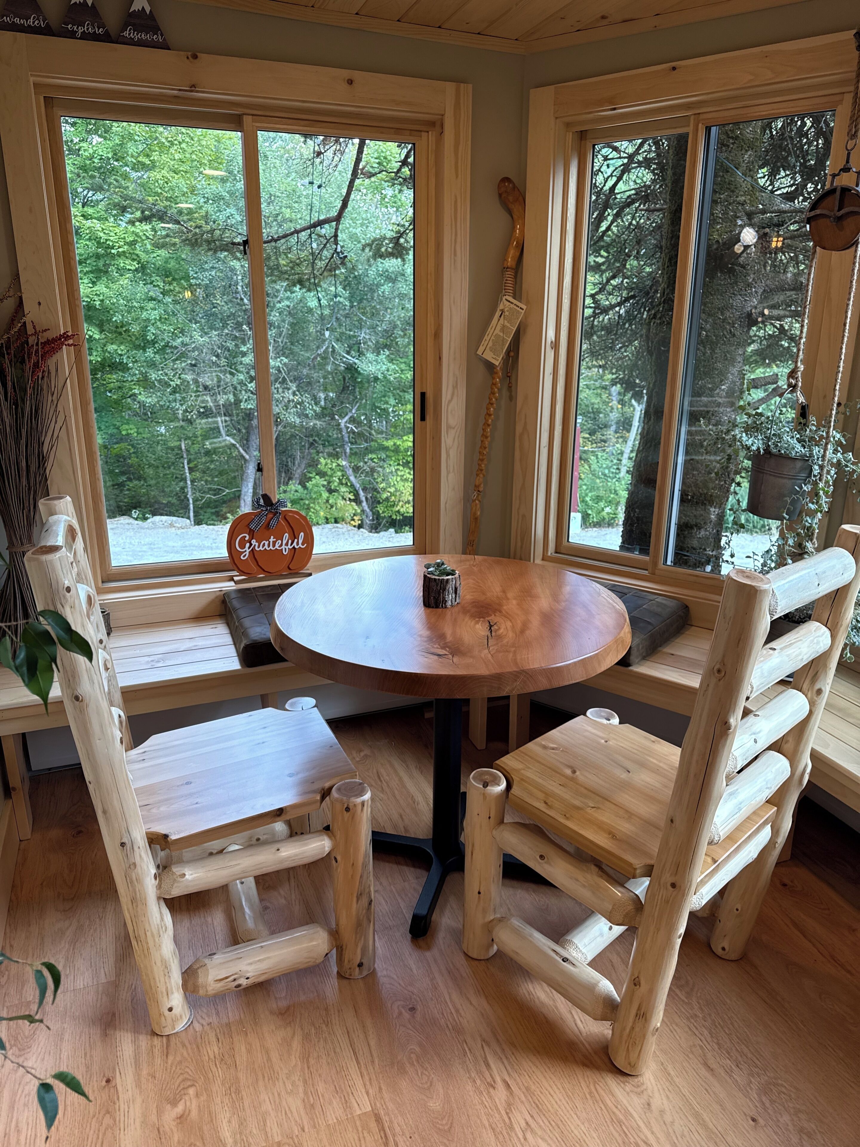 A wooden interior with a round table and benches, large windows with a view of trees, and a hanging wooden sign.