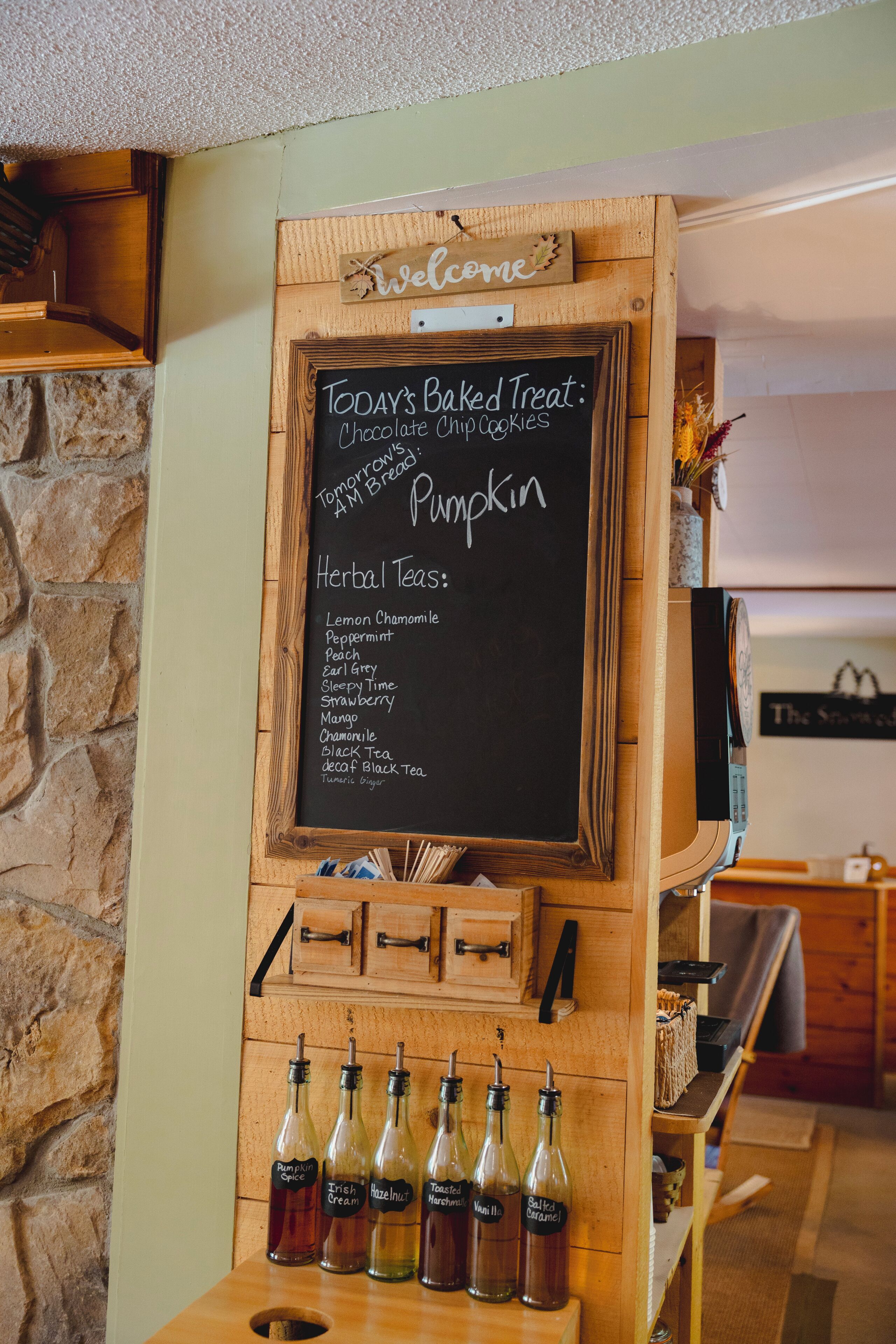 A chalkboard menu listing baked treats and herbal teas, with bottles of various flavors on a wooden table.