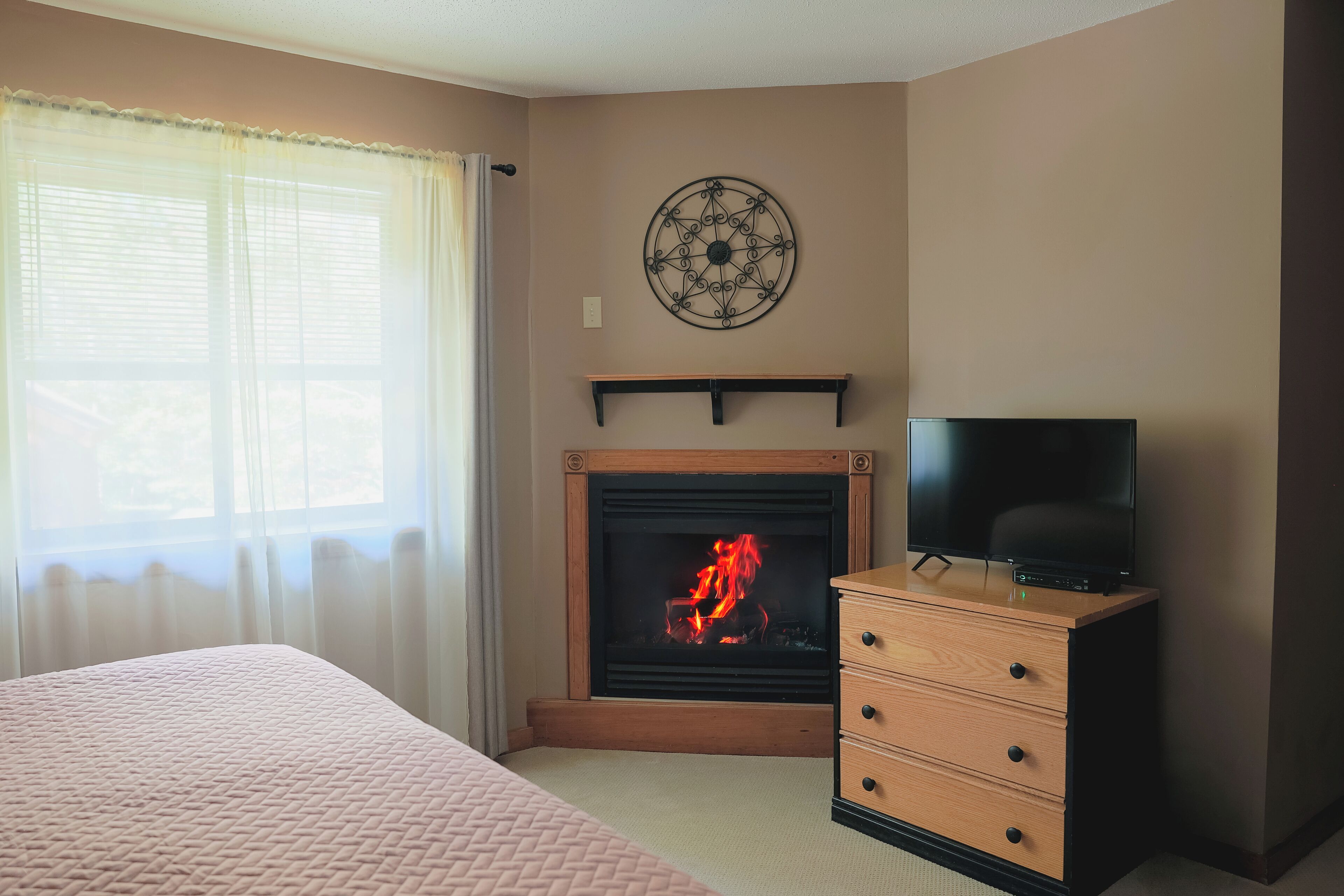 A room with a fireplace, a television on a wooden dresser, and a bed with a pink quilt.