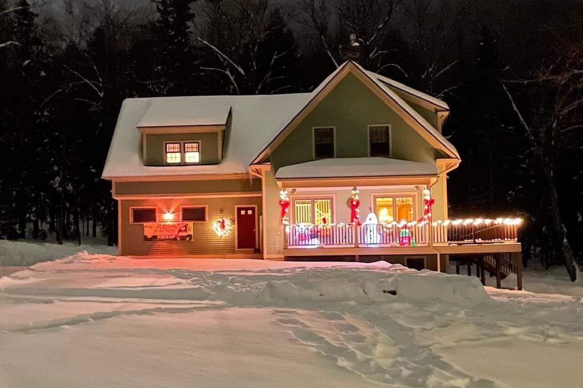 A two-story house with a snow-covered roof and front porch decorated with lights and wreaths.