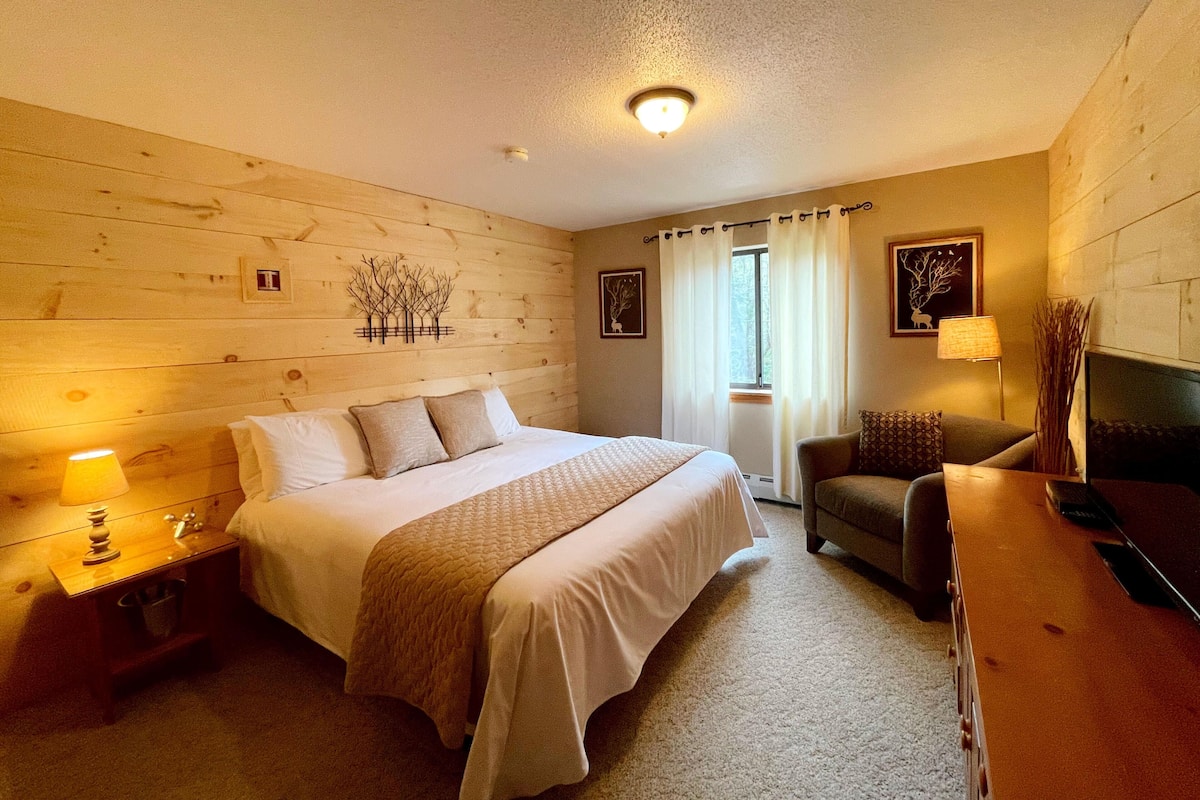 A bedroom with a wooden wall, a bed with a white comforter, a nightstand with a lamp, a chair, and a desk with a lamp.