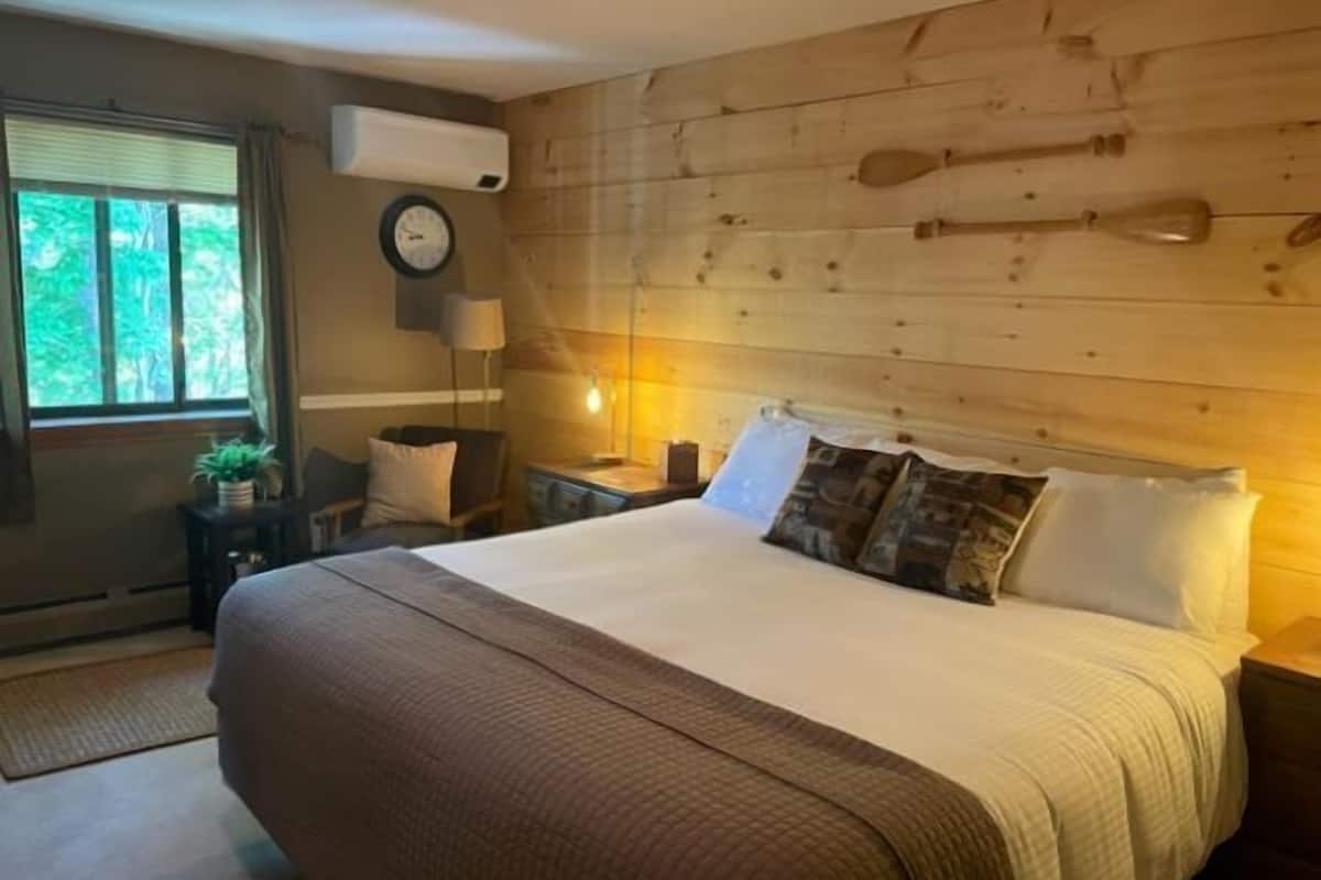 A bedroom with a wooden headboard, a large bed with multiple pillows, a bedside table with a lamp, and a window with curtains.